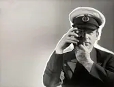 Reklam för filmkamera, Nordiska Kompaniet. Man iklädd skepparhatt och kavaj håller filmkamera framför ögat. Retuscherad bild.