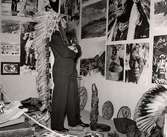 Amerikanska utställningen på varuhuset Nordiska Kompaniet i Stockholm 1938 En man klädd i kostym och med en indiansk fjäderskrud på huvudet betraktar en vägg med uppmonterade foton med indianska motiv. På golvet står artefakter, konsthantverk.