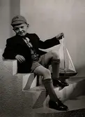 Barnkläder visas på Nordiska Kompaniet 1941. Pojke sitter i trappa iklädd kavaj och kortbyxor, knästrumpor, knytskor och keps. Han håller en segelbåt med ena handen.