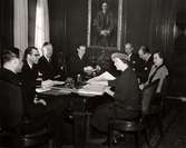 Nordiska Kompaniets budgetkomitté. En grupp män och två kvinnor sitter kring ett ovalt bord. Ovanför ena kortsidan på väggen att mansporträtt, troligen föreställande Josef Sachs.