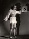 Nordiska Kompaniets kabaré april 1943. En ung kvinna i fransprydda shorts och bolero håller upp en skylt med siffran 6.