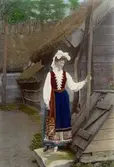 Kvinna i folkdräkt står vid brunn vid ett liggtimrat hus. Klädd i högtidsdräkt från Värend, Småland såsom den tillverkades i rekonstruerad form kring 1900.