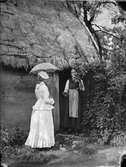 En äldre kvinna står i dörröppningen till en stuga och talar med en elegant klädd dam med uppfällt parasoll. Halland. Troligen förlaga till en målning.