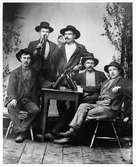 Fem midsommarfirande vägarbetare i ateljén. Männen är arbetsklädda och bär alla hatt, de är grupperade runt ett litet bord, några av dem sitter på pinnstolar och i bakgrunden har lövats med björkris. Männen röker och skålar, några dricker direkt ur flaskan, andra ur glas. Vägbygget gick mellan Lima och Rörbäcksnäs, 1901-1905.