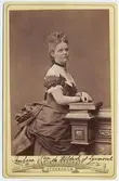 Kabinettsfotografi. Porträtt av prinsessan  Pauline av Waldeck och Pyrmont (1855-1925) iklädd  urringad klänning med turnyr och kort puffärm. Dekorerat dekolletage. Runt halsen bär hon ett band vid vilket ett kors är fäst.1870-tal.