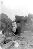 En kvinna med badkappa och mössa sitter på en klippa. På stenarna bredvid henne ligger en väska och klädesplagg, bl.a en korsett.