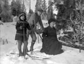 Vinterbild. Gruppfoto av en kvinna som sitter på en sparkstötting och en pojke och en flicka på skidor.