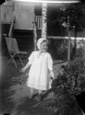 En liten flicka i vit sommarklänning och vit knytmössa på en trädgårdsgång.