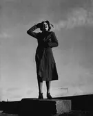 Modell klädd i kappa, snörskor, hatt och handskar, håller handväska under armen. Står utomhus på en sten.