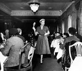 Salén-visning i lunchrummet på Nordiska Kompaniet. Modell klädd i prickig klänning, vita handskar, vit hatt, pärlhalsband och mörka pumps passerar åskådarna. Modellen håller upp en skylt som visar 53SEK.