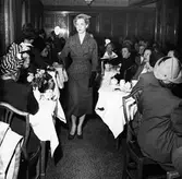 Konfektionsvisning i lunchrummet på Nordiska Kompaniet. Modell passerar publiken klädd i rutig jacka med skärp, kjol, hatt, svarta handskar, vitt pärlhalsband och klackskor. Modellen håller upp en skylt med texten 235 SEK. Publiken sitter vid bord med vita dukar, NK-servis och blommor i vas. De flesta kvinnorna i publiken bär hatt.