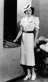 Modell i vit sjömansinspirerad klänning och hatt. Håller väska i handen. Hortensior i bakgrunden.