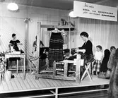 Ebba von Eckermann sitter vid en vävstol och väver Ripsakjolar. På vävstolen hänger en Ripsakjol på en galge. Vid sidan står några barn. I bakgrunden står två skyltdockor klädda i Ripsakjolar.