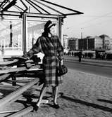 Stilfotografering. Modell i rutig kappa med skärp, lågskor, handväska och hatt. Modellen står i solen och tittar ut över Strömkajen, Stockholm.