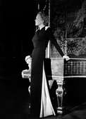 Modell i hellång, svart klänning med lång slits med vitt tyg i sidan. Vita detaljer på axlarna. Modellen lutar sig mot ett piano.