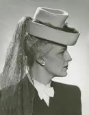 Porträtt av kvinna klädd i hatt med långt dekorband, mörk kavaj, vit blus, vit fluga, samt örhängen.