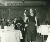 Modevisning på Nordiska Kompaniet, Modell i mörk top, skärp och hellång kjol. Publik sitter vid runda bord.