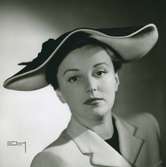 Porträtt av kvinna med hatt i marinblå faille-siden med underbrätte av gul filt. Modell från Gilbert Orcel.
