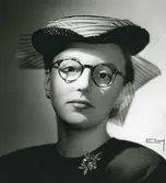 Porträtt av kvinna i glasögon, brosch och liten blå stråhatt med organdimontering i modell av Claude Saint Cyr.