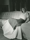 Helporträtt av kvinna i hellång klänning med blockfärger, sittande i en soffa.