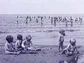 Tbc-sjuka barn inskrivna vid Kustsanatoriet Apelviken på Lilla Apelvikens strand. I förgrunden sitter de yngsta barnen på stranden och i bakgrunden syns en stor grupp äldre barn ute i vattnet. Den lilla flickan på stranden som står upp heter Anna Nilsson och hennes syster Judith sitter bredvid. De kom till sanatoriet från Kalv, då deras mor dött i tbc och de själva var sjuka.