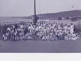 Patienter och personal på stranden vid Lilla Apelviken, Kustsanatoriet. Några av barnpatienterna sitter i sina sängar och två gossar står ovanför gruppen och klamrar sig vid en stolpe. I bakgrunden syns några bodar/badhytter. (Se KSA15 för högre upplösning)