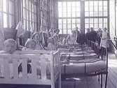 Solveranda på Apelvikens sanatorium med personal och patienter år 1915. I sängarna sitter barnpatienterna och ler mot fotografen. Närmast kameran sitter ett litet barn i spjälsäng och baktill står en grupp ungdomar, bl a en flicka klädd i kappa och vid hatt.