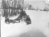 En kvinna sitter med tre barn i en snöbacke. Hon är i färd med att kasta en snöboll, en flicka sitter på en sparkstötting och en annan ligger ned.
