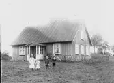 Fyra barn står med en höna framför ett omålat bostadshus med brant, bandplåt på taket. Fönstren är ovanligt stora med korspost. Möjligen är det en skola. Gaveln omgärdas av ett enkelt staket, troligen ämnat att skydda växter innanför det mot lösgående djur.