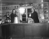 Interiör från en affär av outgrundligt slag där två kvinnor står bakom disken. Till höger ett glasat skåp med metallföremål såsom kannor, urnor, ljusstakar etc. De öppna hyllorna till vänster är tomma. Ena kvinnan stöttar armbågen på en hink och där finns en ställning med inslagspapper.