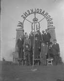 En grupp män framför något slags monument där en båge med kunglig symbol, kungakrona och texten 