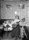 En ung kvinna sitter och spelar gitarr. Bordet bredvid är fullt av små inramade fotografier och på piedestalen sitter ett leksaksdjur.