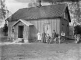 Famlij framför enkelhus i Hede med husdjur och trattgrammofon