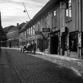 En man bredvid en häst med vagn går på Smedjegatan i Jönköping, förbi Nya garnboden, 8:ans resandevåning,  Ericsons konditori och en tobaksaffär .