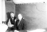 En man med pipa (1) och fotograf Gustav Andersson (2) sitter på sängen i ett rum med blommiga tapeter, på väggen bakom dem hänger ett fickur på en krok.
