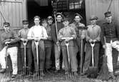 Tio män klädda i uniformsmössor och tre av dem i uniformsjackor, står framför två öppna trädörrar. De är utrustade med en spade, en kvast, två borstar samt fyra högafflar.