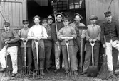 Tio män klädda i uniformsmössor och tre av dem i uniformsjackor, står framför två öppna trädörrar. De är utrustade med en spade, en kvast, två borstar samt fyra högafflar.