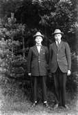 Två män, Gustav Andersson (2), står på en väg i skogen, klädda i kostym och hatt.