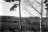 Ett bostadshus i trä ligger omgiven av åkrar, i bakgrunden syns skogsklädda höjder. I förgrunden står två tallar och en avlövad björk.