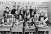 Fröken R. Sylvén med 1:a klass-elever på Centralskolan 1943-44. Flickor sittandes i bänkar. Flickor och pojkar ståendes bakom. Teckningar på väggen. Med på bild finns, sittandes till höger, Berit Gustafsson (född Larsson 1936).