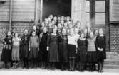 Flickor i 7:e klassen, stående framför Trädgårdsskolans (Kommunala mellanskolans) port.
Längst bak står lärarinnan fröken Thorsson och överlärare Ernst Lindstrand.