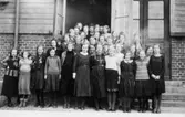 Flickor i 7:e klassen, stående framför Trädgårdsskolans (Kommunala mellanskolans) port.
Längst bak står lärarinnan fröken Thorsson och överlärare Ernst Lindstrand.