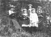 Troligen en lärare/guvernant med barn/ungdomar, sittandes och ståendes utomhus i en slänt vid en träddunge.
Från början av 1900-talet.
Bleknat fotografi. Okänd plats.