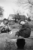 Marie-Louise Lindholm (gift Carlsson) och Lisbeth leker med spade och mugg i trädgården. Flickorna leker på Görjekullen (längst upp på nuvarande Åldermansgatan). I bakgrunden ses Roten K 18, 1950-tal.