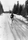 Lantbrevbärare, postiljon Erik Bergman (cykelåkande) i
Delsbo.  April 1956.

Brevbäringsturen är 2 ½ mil lång och omfattar nästan 300 hushåll. Postväskan väger ofta 30-40 kg.