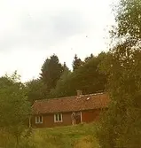 Ett rödmålat bostadshus, ett och ett halvt plan, som ligger på en kulle. Ort och årtal är okänt.