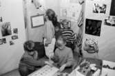 Tre barn som pysslar med något på golvet. Några barn tittar på saker som är uppsatta på en vägg. Utställningsvernissage av och om Katrinebergs daghem på Mölndals museum 1993-09-10.