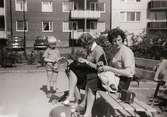 Två kvinnor, Lisbert Andersson och Kyllan Lageklöv, sitter på en bänk på Lerbrännaregatans innegård, 1960-tal. De passar på att handarbeta när barnen leker. Till vänster står ett barn. Kvinnorna är bekanta till fotografen Astrid. Familjen Garthman flyttade till Lerbrännargatan 1C, 1960-09-01.