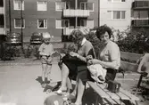 Två kvinnor, Lisbert Andersson och Kyllan Lageklöv, sitter på en bänk på Lerbrännaregatans innegård, 1960-tal. De passar på att handarbeta när barnen leker. Till vänster står ett barn. Kvinnorna är bekanta till fotografen Astrid. Familjen Garthman flyttade till Lerbrännargatan 1C, 1960-09-01.