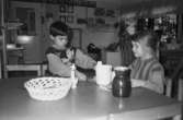 Två dagisbarn (en pojke och en flicka) som fikar sittandes vid ett bord. Katrinebergs daghem 1992-93.